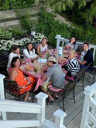 Eight adults gathered around a red-checkered picnic table on a backyard wooden deck, enjoying a casual summer meal with plates, drinks and daisies in the surrounding garden.