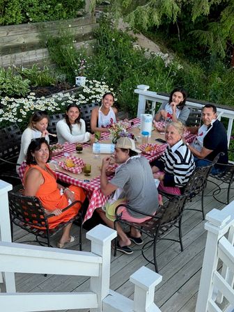 Eight adults gathered around a red-checkered picnic table on a backyard wooden deck, enjoying a casual summer meal with plates, drinks and daisies in the surrounding garden.