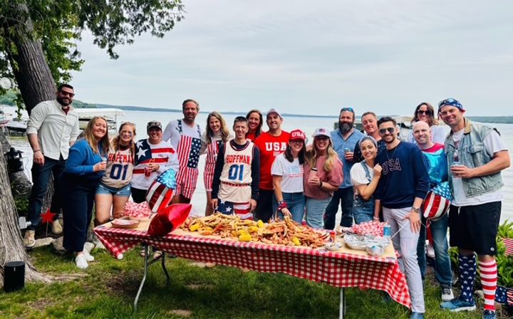 Lakeside group of adults in patriotic red, white and blue outfits posing at a summer picnic by the water, a checkered table piled with a seafood boil of crab legs, corn and sides.