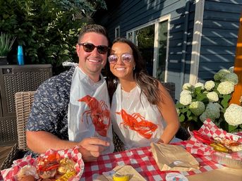 Two smiling people in sunglasses wearing lobster bibs at an outdoor patio seafood boil — red-checkered tablecloth with corn, shellfish and dipping sauce, white hydrangeas and blue house siding in background.