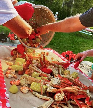 Hands dumping a colorful seafood boil — crab legs, shrimp, corn, sausage and potatoes — onto a paper-covered picnic table at a backyard summer gathering