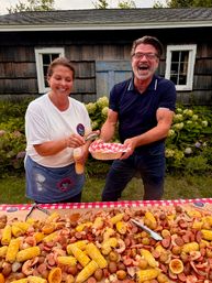 Two smiling people serving a backyard seafood boil—corn on the cob, shrimp, sausage and potatoes—on a red-checkered picnic table in front of a rustic wooden shed.