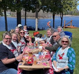 Smiling group enjoying a lakeside lobster picnic under trees on a sunny day, red-checkered table, lobster bibs and a dock in the background.