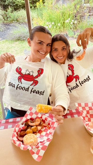 Two smiling women at an outdoor seafood boil wearing cartoon-crab “seafood feast” bibs, sharing shrimp, corn on the cob and potatoes in red-checkered baskets.