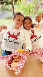 Two smiling women at an outdoor seafood boil wearing cartoon-crab “seafood feast” bibs, sharing shrimp, corn on the cob and potatoes in red-checkered baskets.