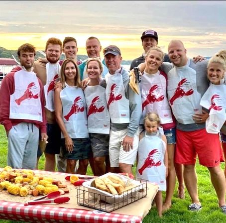 Group of family and friends wearing lobster bibs at a lakeside sunset seafood boil — outdoor summer picnic with corn on the cob, potatoes and bread on a red-check tablecloth