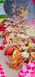 Outdoor seafood boil being poured from a metal colander onto a paper-lined table — crab legs, crawfish, shrimp, corn on the cob, sausage slices and baby potatoes on red checkered paper.