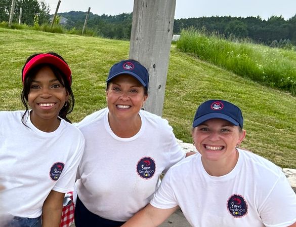 Three smiling people in matching white t-shirts and navy/red caps posing under a wooden post at a grassy outdoor summer event near rolling hills.