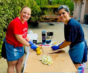 Two smiling people in denim aprons and blue gloves prepping lemons and onion peels on kraft paper at a backyard patio/driveway table, with a cooler, thermos and portable speaker nearby — outdoor meal prep vibe.