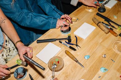 Close-up of an artisan jewelry workshop bench with hands at work amid hammers, mallet, pliers, chisels, polishing pads, a steel mandrel, turquoise cabochons and ring settings on a light wooden table.