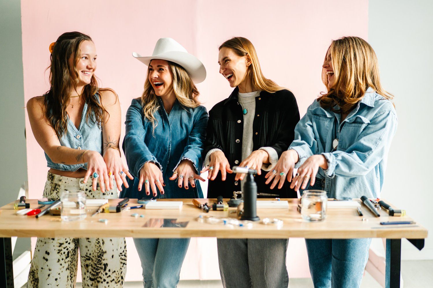 Four smiling women in a bright craft studio standing at a workbench, holding out hands to show handmade rings and jewelry tools, pliers, torch and jars on the table; casual denim outfits and a white cowboy hat add playful flair.
