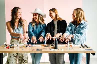 Four smiling women in a bright craft studio standing at a workbench, holding out hands to show handmade rings and jewelry tools, pliers, torch and jars on the table; casual denim outfits and a white cowboy hat add playful flair.