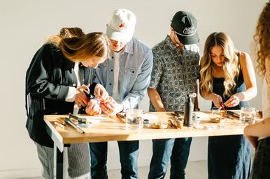 Small group of people standing at a wooden table in a bright studio, focused on a hands-on jewelry-making workshop with pliers, small tools, and a tabletop torch.