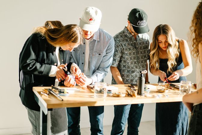 Small group of people standing at a wooden table in a bright studio, focused on a hands-on jewelry-making workshop with pliers, small tools, and a tabletop torch.