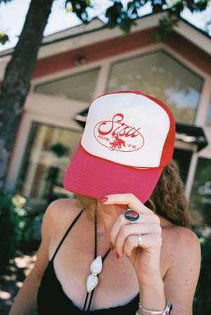 Person hiding face under red-and-white trucker hat with cowboy graphic and 'AUS TX' text, wearing a black bikini top, silver stone necklace and rings — summer Austin, Texas street-style shot.