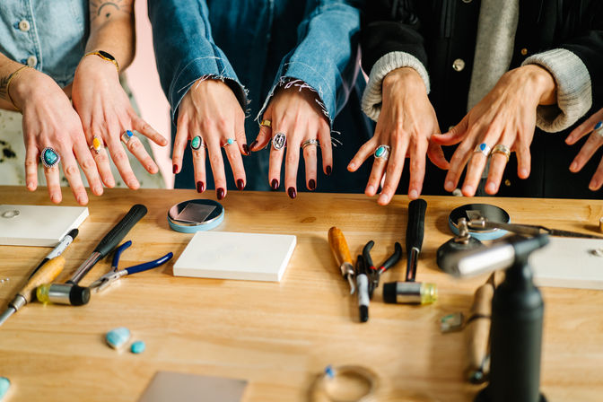 Several hands wearing colorful turquoise and silver rings stretched over a wooden workbench in a jewelry-making studio, surrounded by hammers, pliers, mallet, tiles and small gemstone pieces, ready to craft.
