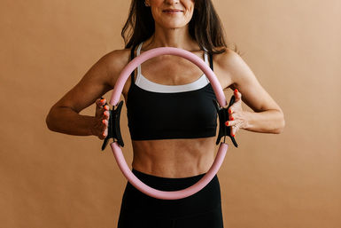 Fit person in a black sports bra holding a pink Pilates ring at chest level for a core-strengthening fitness workout against a neutral tan background.