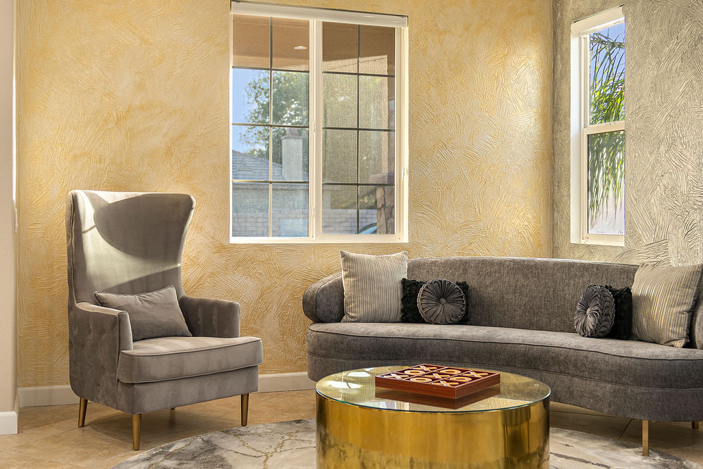 Sunlit modern living room interior with a gray velvet wingback armchair, curved gray sofa with decorative pillows, round gold glass-top coffee table and textured beige plaster walls by two windows.