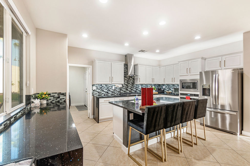 Sunlit modern kitchen with white cabinets, mosaic tile backsplash, stainless steel appliances, black quartz center island with three velvet bar stools and beige tile floor