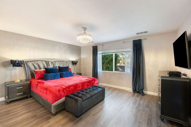 Contemporary master bedroom with bold red velvet bedspread, tufted gray headboard, navy accent pillows, crystal chandelier, dark nightstands and bench, hardwood floors, large window with navy curtains and wall-mounted TV.