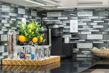 Modern kitchen countertop with a black single-serve coffee maker, reflective glass-tile backsplash, tray of glassware and coffee pods, and a potted plant with bright yellow decorative lemons.