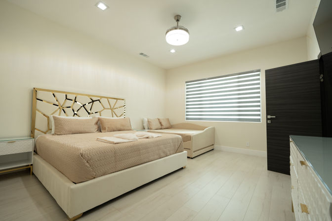 Bright modern master bedroom with beige upholstered king bed, geometric gold-accent headboard, matching daybed, striped zebra window shade, light wood floors and round ceiling light