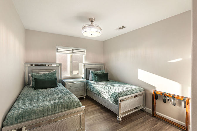 Sunlit modern twin bedroom with metallic silver-framed beds, teal quilted bedding and matching pillows, mirrored nightstand between beds, hardwood floors and neutral walls.
