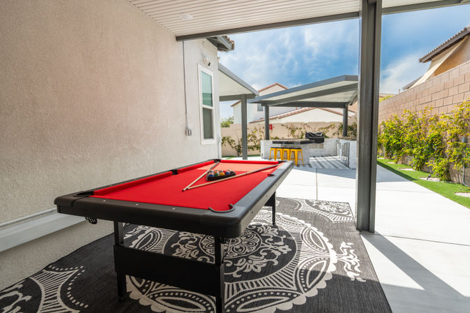 Covered backyard patio featuring a red-felt pool table on a patterned outdoor rug with cues and racked balls, opening to a modern outdoor kitchen and bar with yellow stools under a pergola and a landscaped privacy wall.