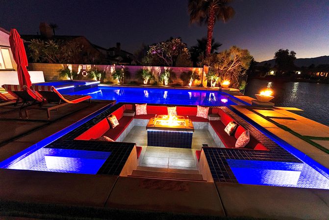 Luxury waterfront backyard at night featuring a glowing blue pool, sunken red-cushioned conversation pit with a central fire pit, tiled steps, lounge chairs, palm trees and fire bowls reflecting on a calm lake