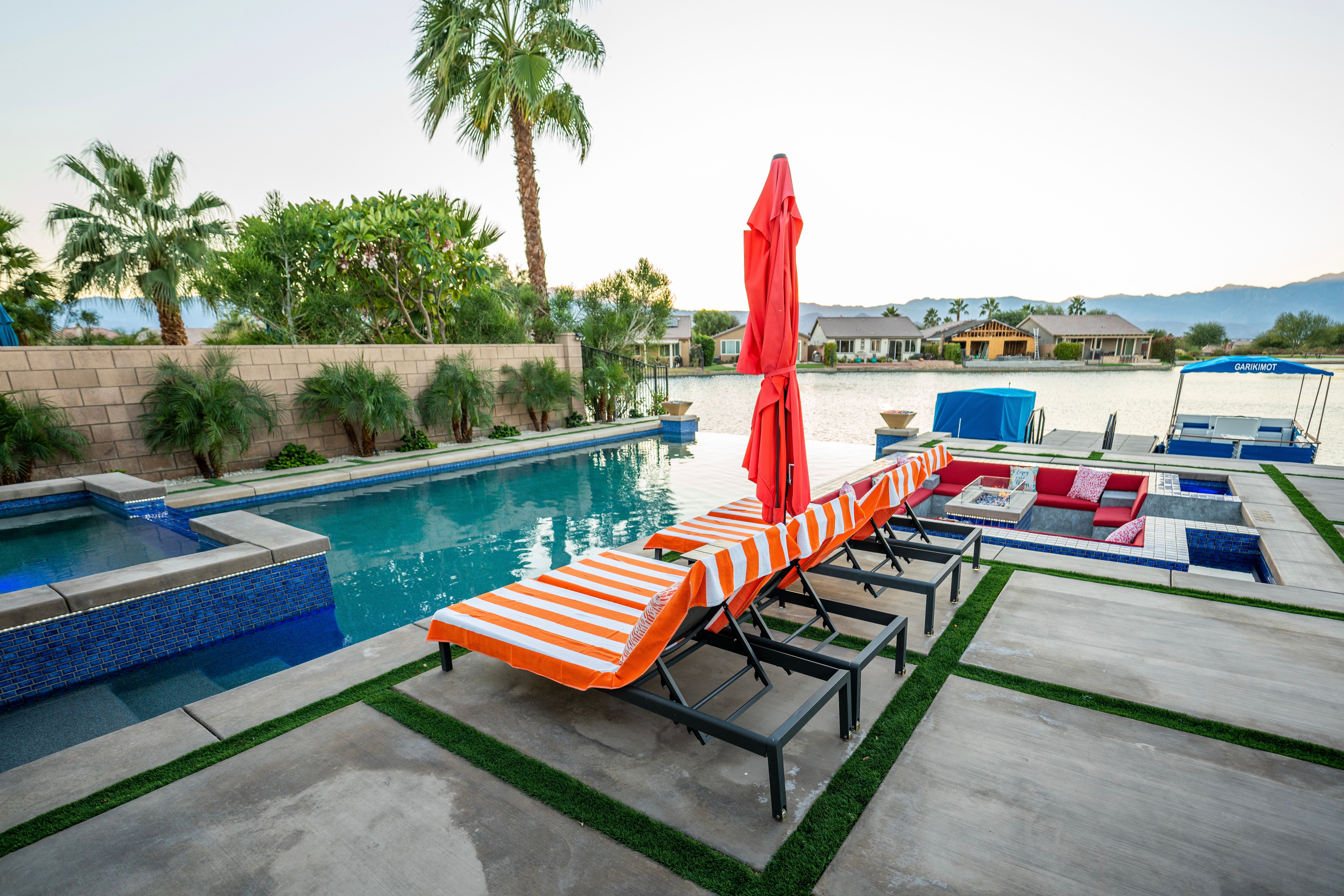 Lakefront luxury backyard with pool, orange-striped lounge chairs and red umbrella, sunken sofa firepit area, palm trees and mountain backdrop