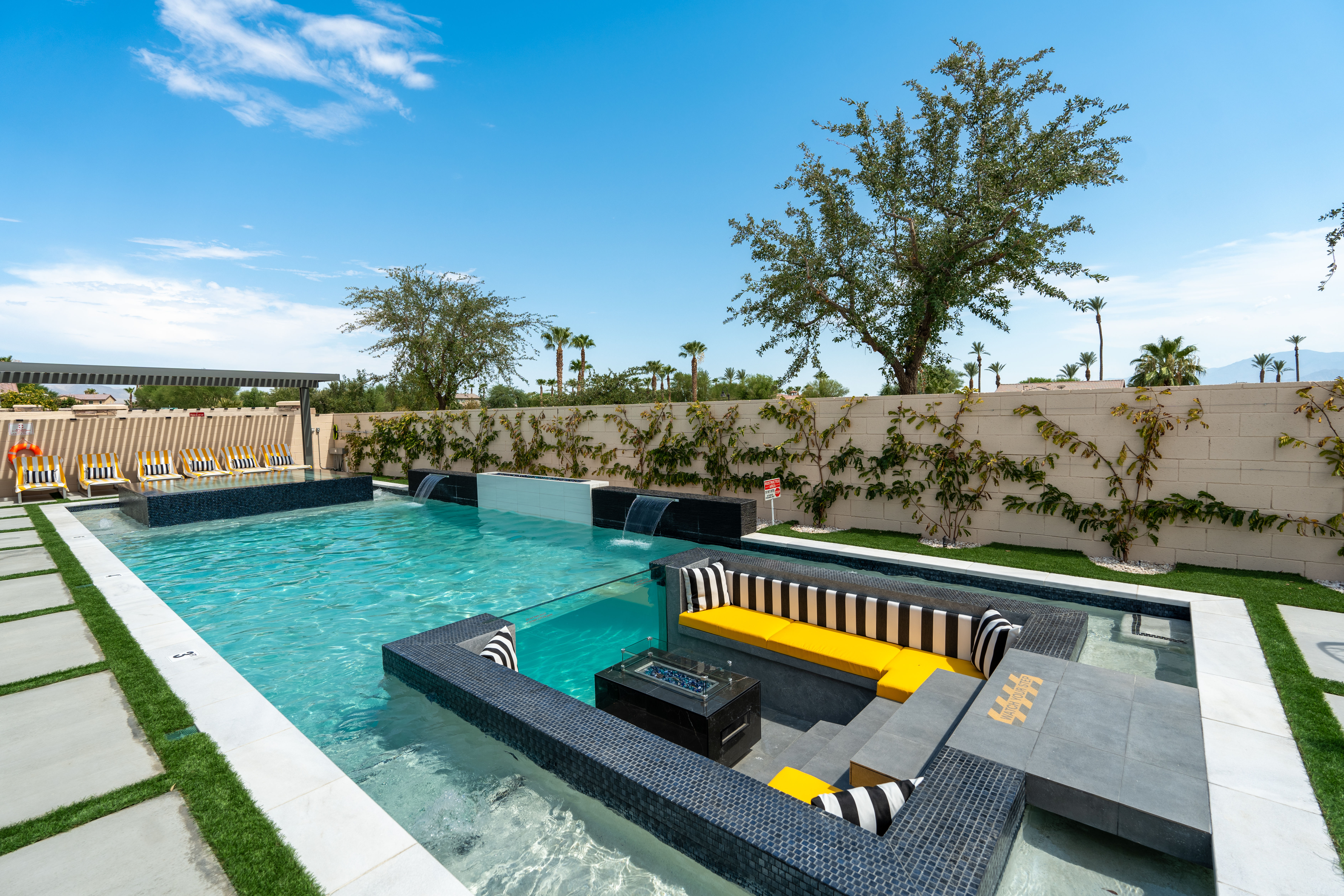 Modern luxury backyard pool with built-in sunken seating and yellow-striped cushions, cascading water features, poolside lounge chairs, palm trees and bright blue sky