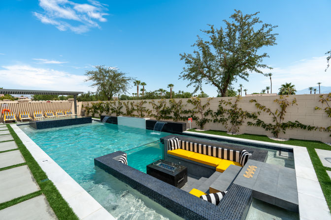 Modern luxury backyard pool with built-in sunken seating and yellow-striped cushions, cascading water features, poolside lounge chairs, palm trees and bright blue sky
