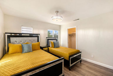 Cheery sunlit modern bedroom with two beds dressed in yellow quilted bedding, mirrored black frames, hardwood floors, window, and small open closet.