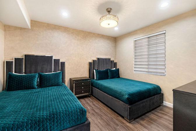 Stylish modern guest bedroom with two teal quilted beds, velvet paneled headboards, dark nightstand, wood-look flooring and neutral walls