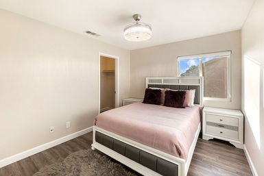 Cozy modern residential bedroom with queen bed in pink bedding, plush dark pillows, faux-leather headboard, white nightstands, wood-look flooring, shag rug, walk-in closet and ceiling light.