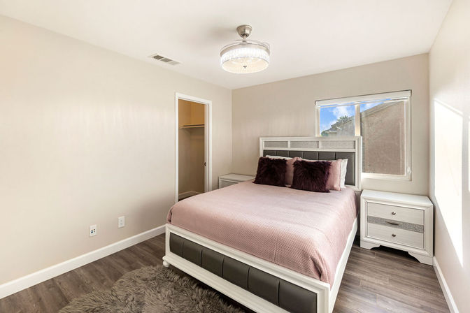 Cozy modern residential bedroom with queen bed in pink bedding, plush dark pillows, faux-leather headboard, white nightstands, wood-look flooring, shag rug, walk-in closet and ceiling light.