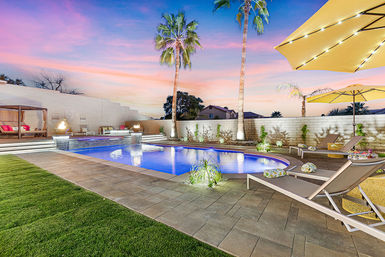 Twilight backyard pool with lit rectangular pool, tall palm trees, cabana, fire bowls and lounge chairs under yellow umbrellas on a paved patio at sunset