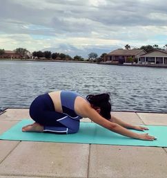 Person in child's pose on a teal yoga mat on a lakeside concrete dock, calm water and waterfront homes under a cloudy sky — relaxed lakeside yoga scene.