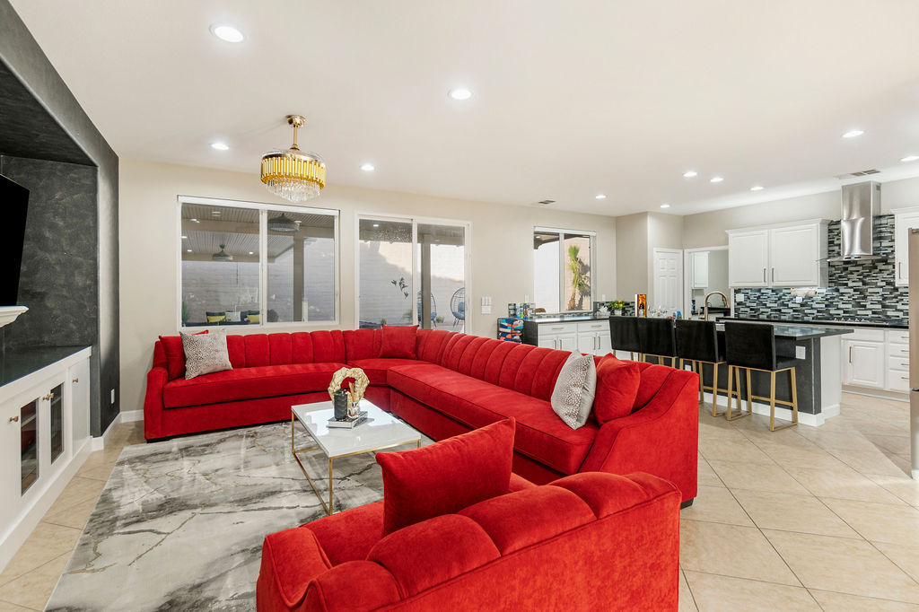 Open-concept modern living room and kitchen with a bold red U-shaped sectional, gold chandelier, marble-effect rug, tile floors, and a black-stool breakfast bar.
