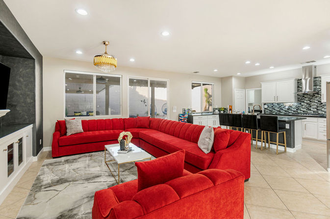Open-concept modern living room and kitchen with a bold red U-shaped sectional, gold chandelier, marble-effect rug, tile floors, and a black-stool breakfast bar.