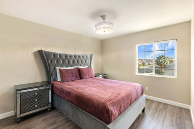 Stylish bright bedroom with tufted gray headboard, burgundy quilted bed, mirrored nightstands, and a window view of a sunny suburban palm-lined street.