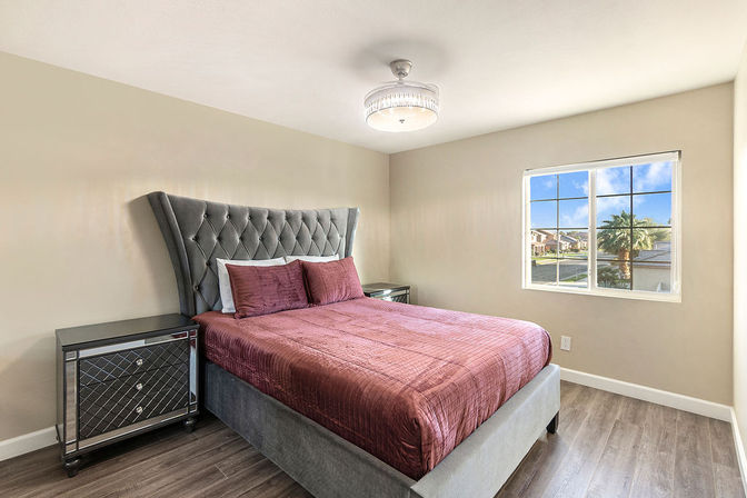 Stylish bright bedroom with tufted gray headboard, burgundy quilted bed, mirrored nightstands, and a window view of a sunny suburban palm-lined street.