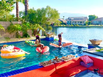 Lakefront backyard pool scene with people on colorful inflatable floats in a turquoise pool, red patio seating with trays of snacks, palm trees, houses and distant mountains under a sunny sky.