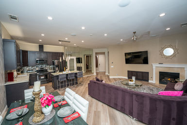 Modern open-concept living room and kitchen in a residential home with a curved purple sectional, round coffee table and fireplace, wall-mounted TV, dark wood cabinets and stainless appliances, breakfast island with barstools, dining table set, and light wood floors.