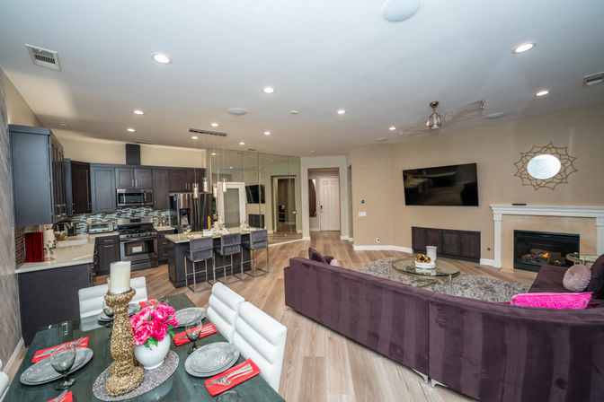 Modern open-concept living room and kitchen in a residential home with a curved purple sectional, round coffee table and fireplace, wall-mounted TV, dark wood cabinets and stainless appliances, breakfast island with barstools, dining table set, and light wood floors.