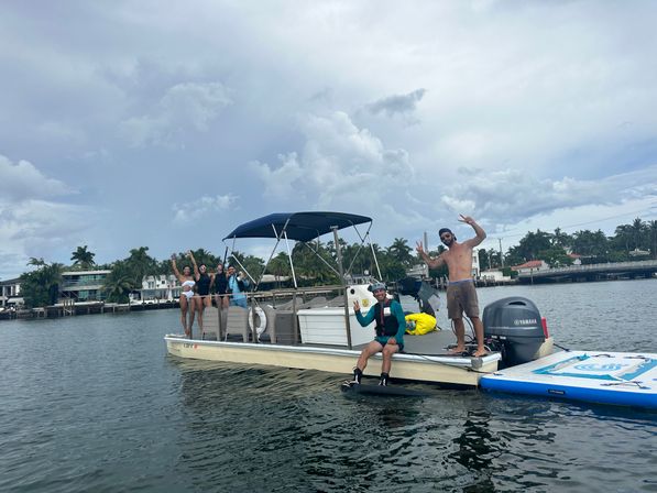 Group of friends on a pontoon boat in a tropical waterfront canal with palm trees and houses, paddleboard and wakeboard at the stern and a cloudy sky overhead.