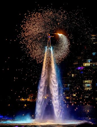 Flyboard performer propelled by twin water jets at night, swinging a flaming spark wand that sprays golden pyrotechnic sparks into a halo above a lit waterfront and city buildings.