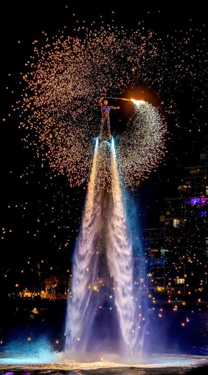 Flyboard performer propelled by twin water jets at night, swinging a flaming spark wand that sprays golden pyrotechnic sparks into a halo above a lit waterfront and city buildings.