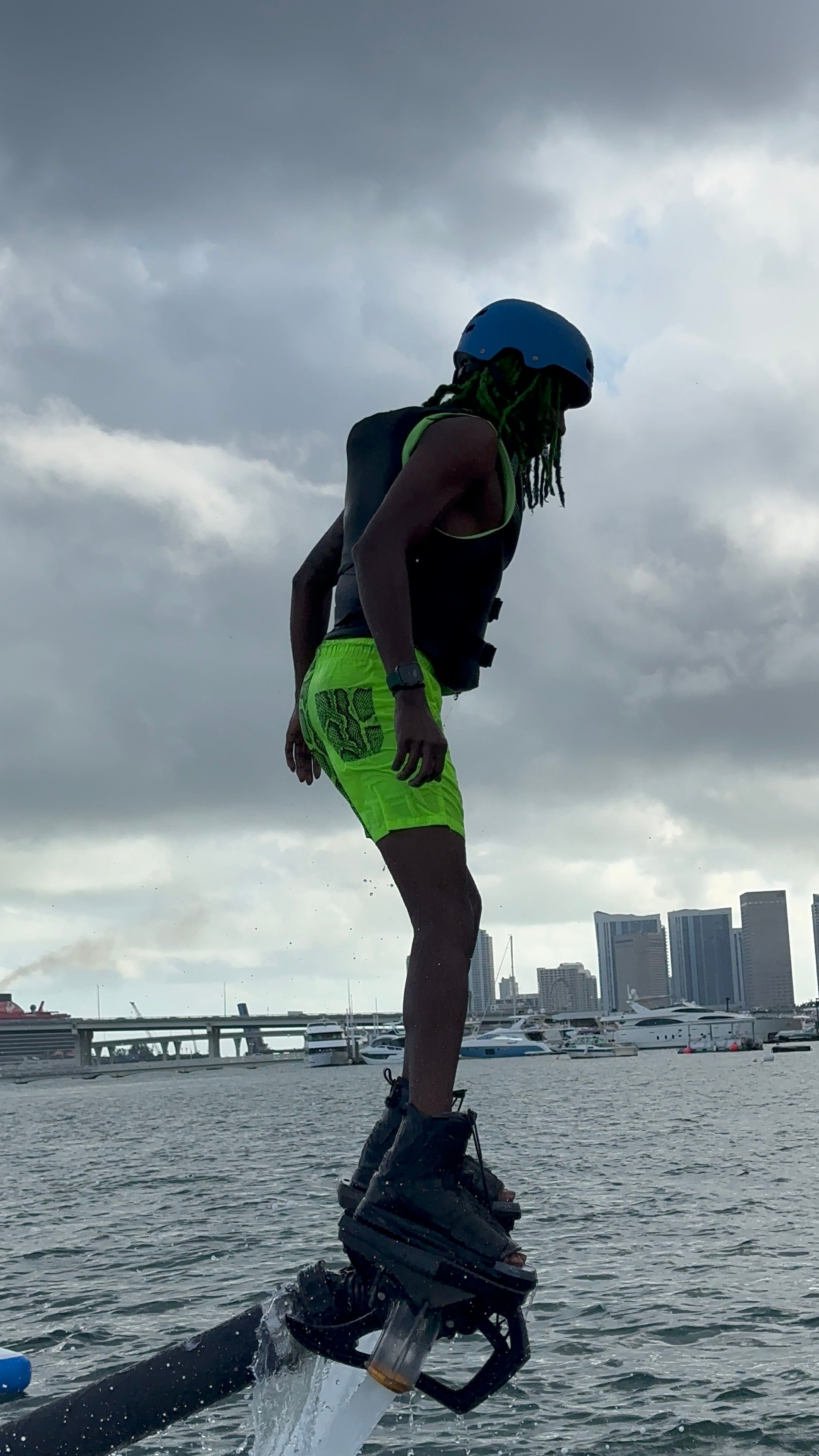 Flyboard rider in a blue helmet and neon green shorts hovering above an urban marina with yachts and a cloudy city skyline
