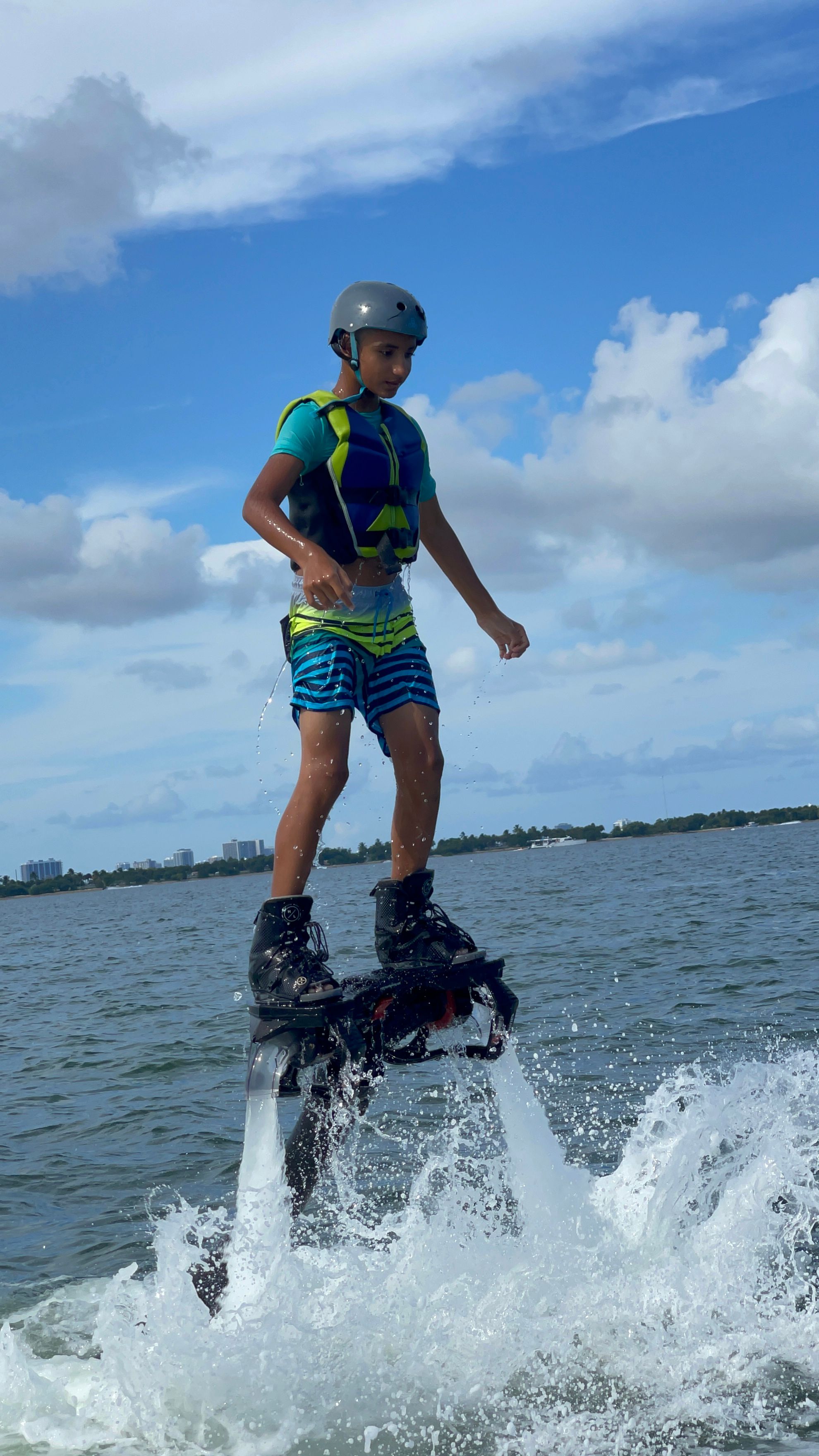 Young person flyboarding over a coastal bay, wearing a helmet and life vest as powerful water jets spray beneath, blue sky and distant shoreline in the background